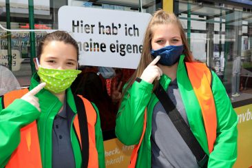 zwei Mädchen tragen eine Stoffmaske über Mund und Nase und HAndschuhe, um auf die Maskenpflicht in Bussen und Bahnen aufmerksam zu machen.