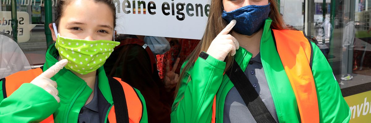 zwei Mädchen tragen eine Stoffmaske über Mund und Nase und HAndschuhe, um auf die Maskenpflicht in Bussen und Bahnen aufmerksam zu machen.
