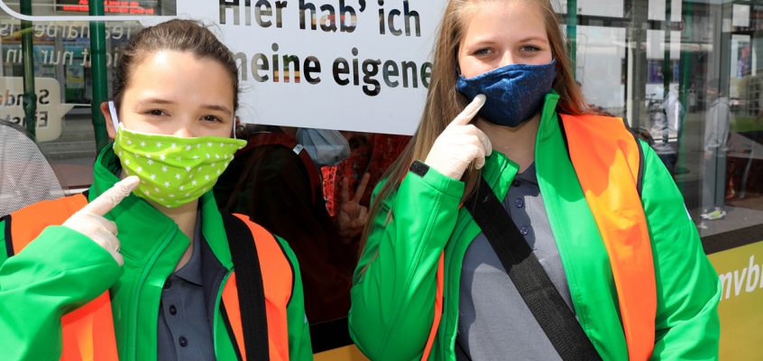 zwei Mädchen tragen eine Stoffmaske über Mund und Nase und HAndschuhe, um auf die Maskenpflicht in Bussen und Bahnen aufmerksam zu machen.