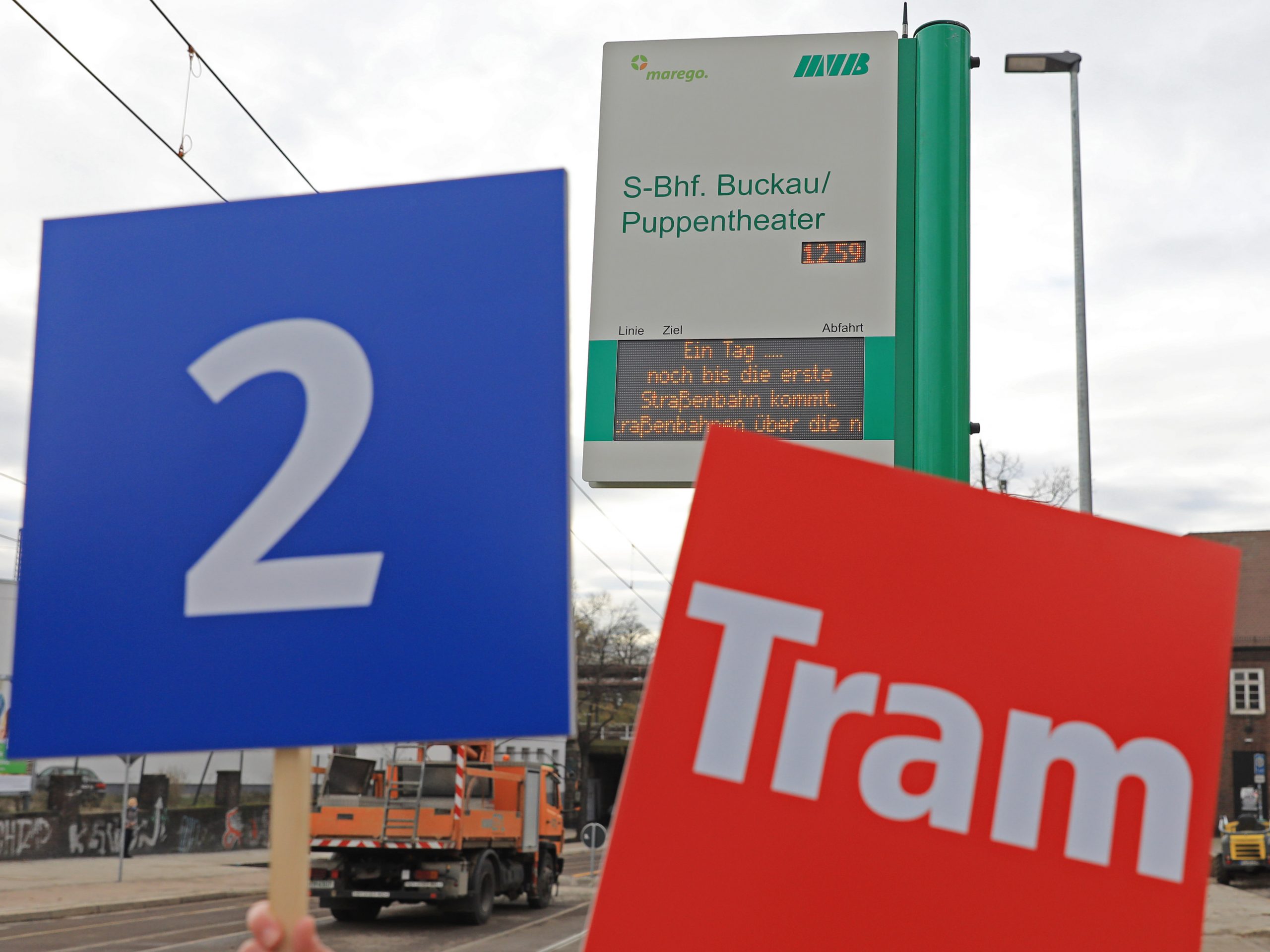 Zur Einweihung der neuen Strecke Puppentheater Bahnhof Buckau Warschauer Str. werden Schilder mit der Nummer 2 und dem Tram Logo vor der Fahrgastinformation an der Haltestelle S-Bhf. Buckau/Puppentheater in die Kamera gehalten.