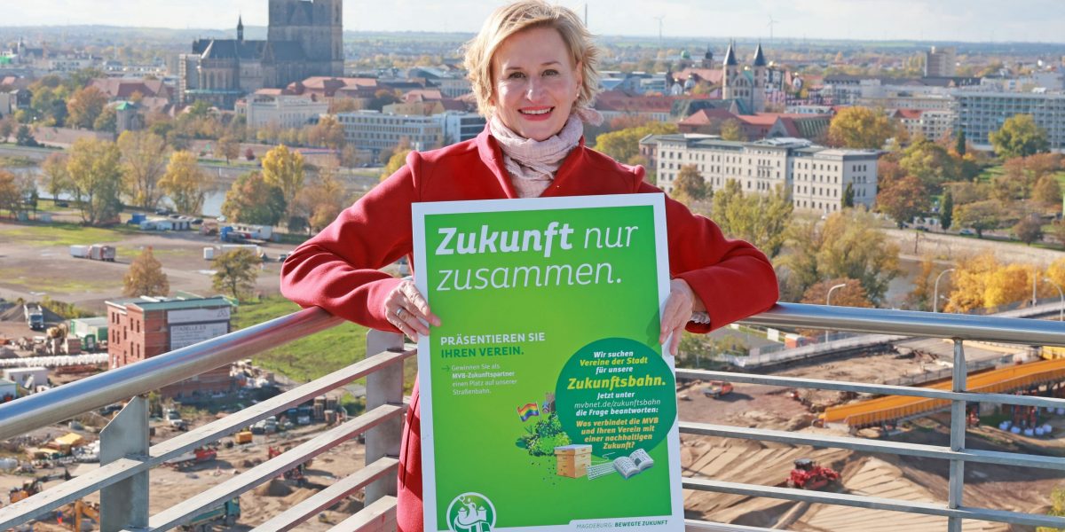 Abteilungsleiterin vom Bereich Marketing, Cornelia Muhl-Hünicke, mit einem Schild zur Zukunftsbahn. Im Hintergrund ist die Skyline von Magdeburg zu sehen.