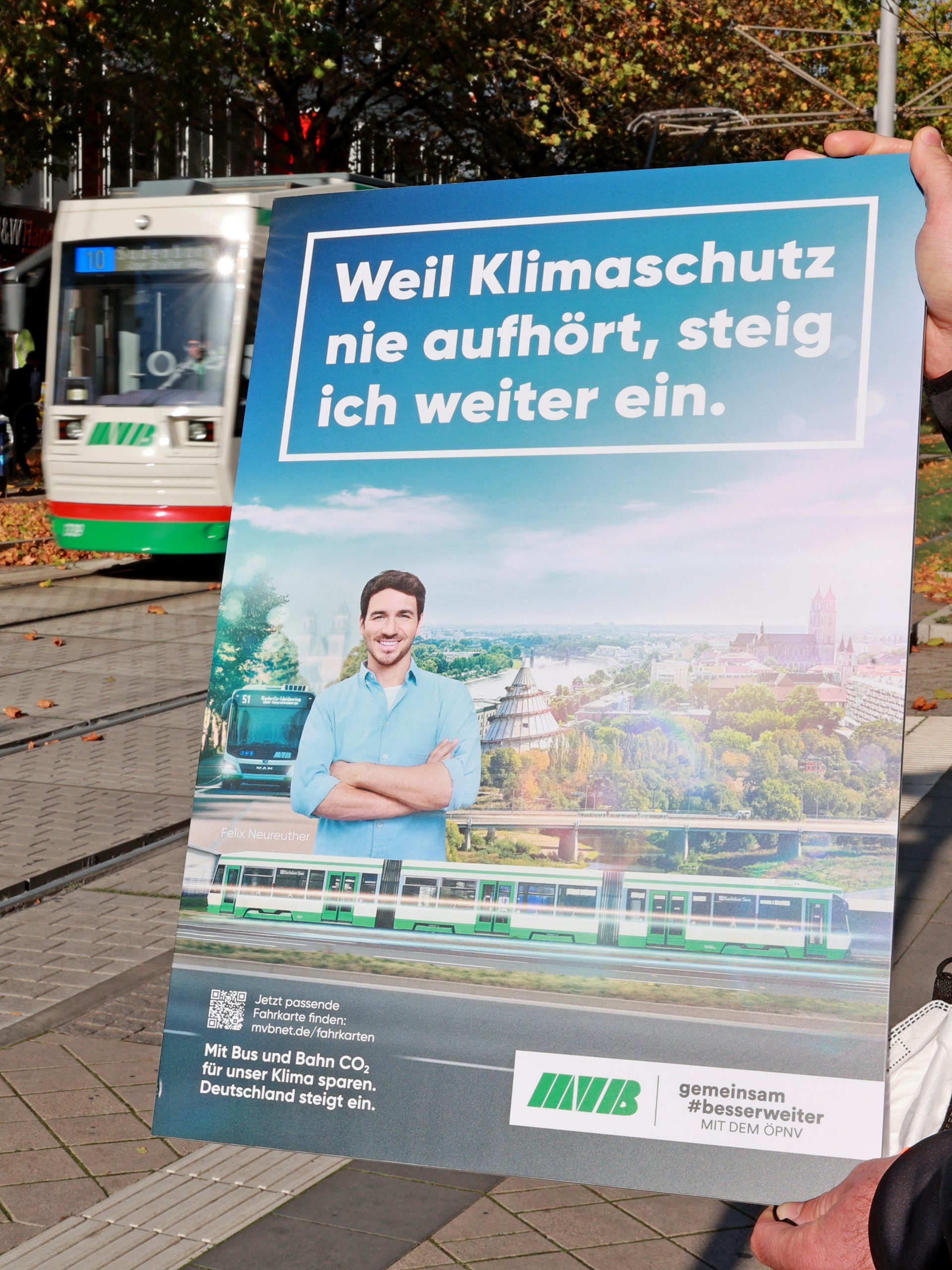 Jemand hält ein Plakat zu einer Klimaschutzaktion hoch Im Hintergrund ist eine Straßenbahn zu sehen.