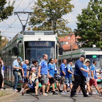 blau weiß gekleidete Fußballfans des 1. FC Magdeburgs überqueren vor einer Straßenbahn die Straße an der Haltestelle Arenen
