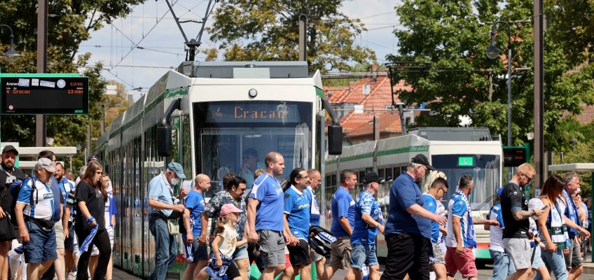 blau weiß gekleidete Fußballfans des 1. FC Magdeburgs überqueren vor einer Straßenbahn die Straße an der Haltestelle Arenen