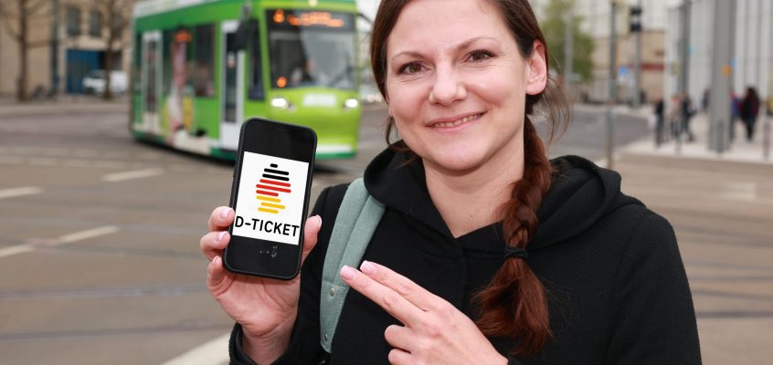 Eine Frau mit dem Smartphone in der Hand, auf dem das D-Ticket-Logo zu sehen ist. Straßenbahnen sind im Hintergrund zu sehen.