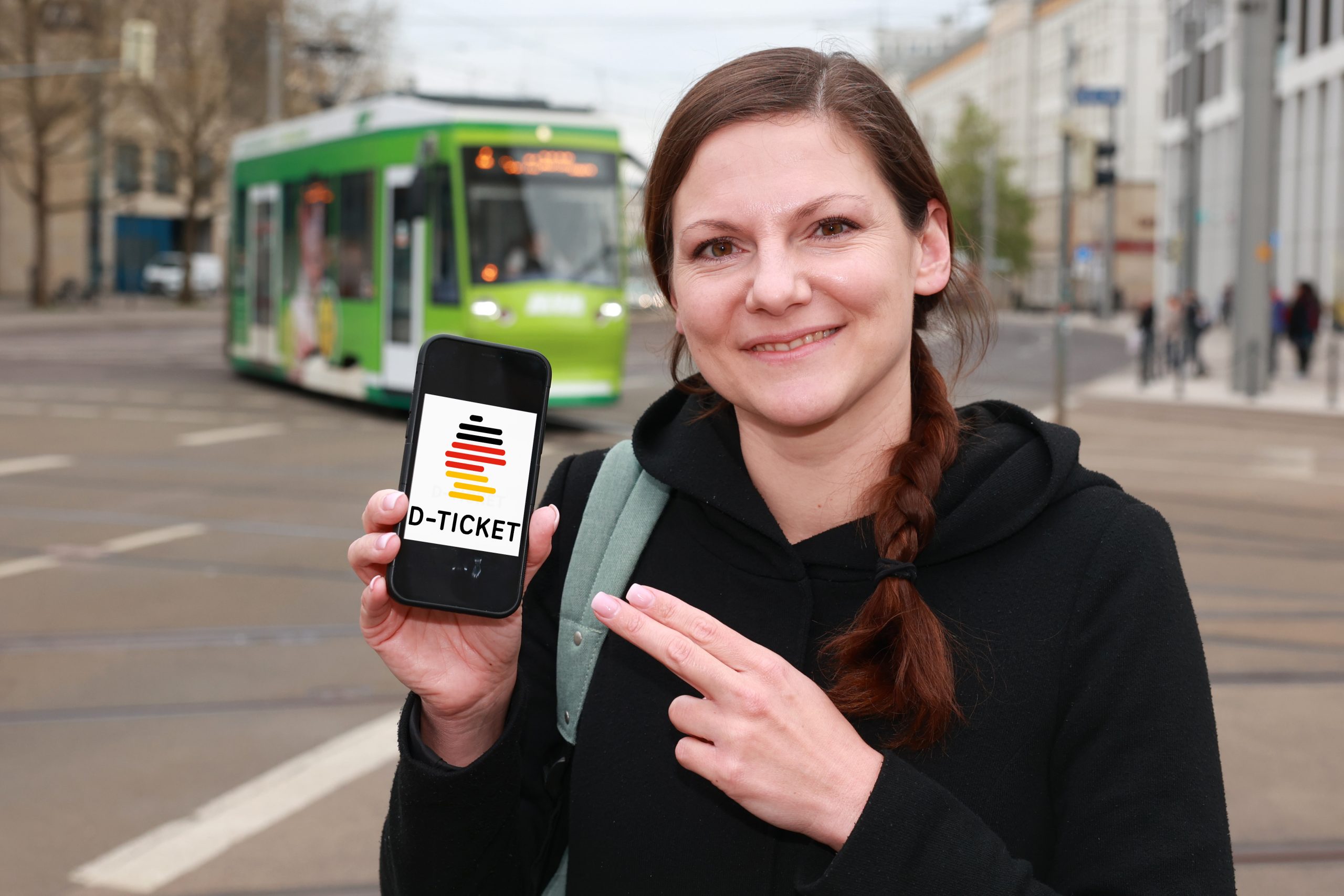 Eine Frau mit dem Smartphone in der Hand, auf dem das D-Ticket-Logo zu sehen ist. Straßenbahnen sind im Hintergrund zu sehen.