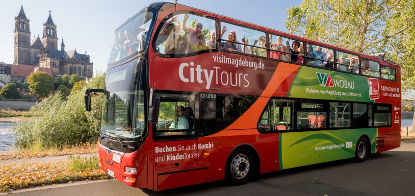 Winkende und glückliche Menschen auf der oberen Etage des Doppeldecker Busses von City Tours im Stadtpark. Im Hintergrund sind der Dom und die Elbe zu sehen.