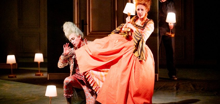 Werbefoto aus dem Theaterstück Die Hochzeit des Figaro. Zwei Personen in barocker Kleidung sind auf der Bühne, eine dritte versteckt sich im Hintergrund,