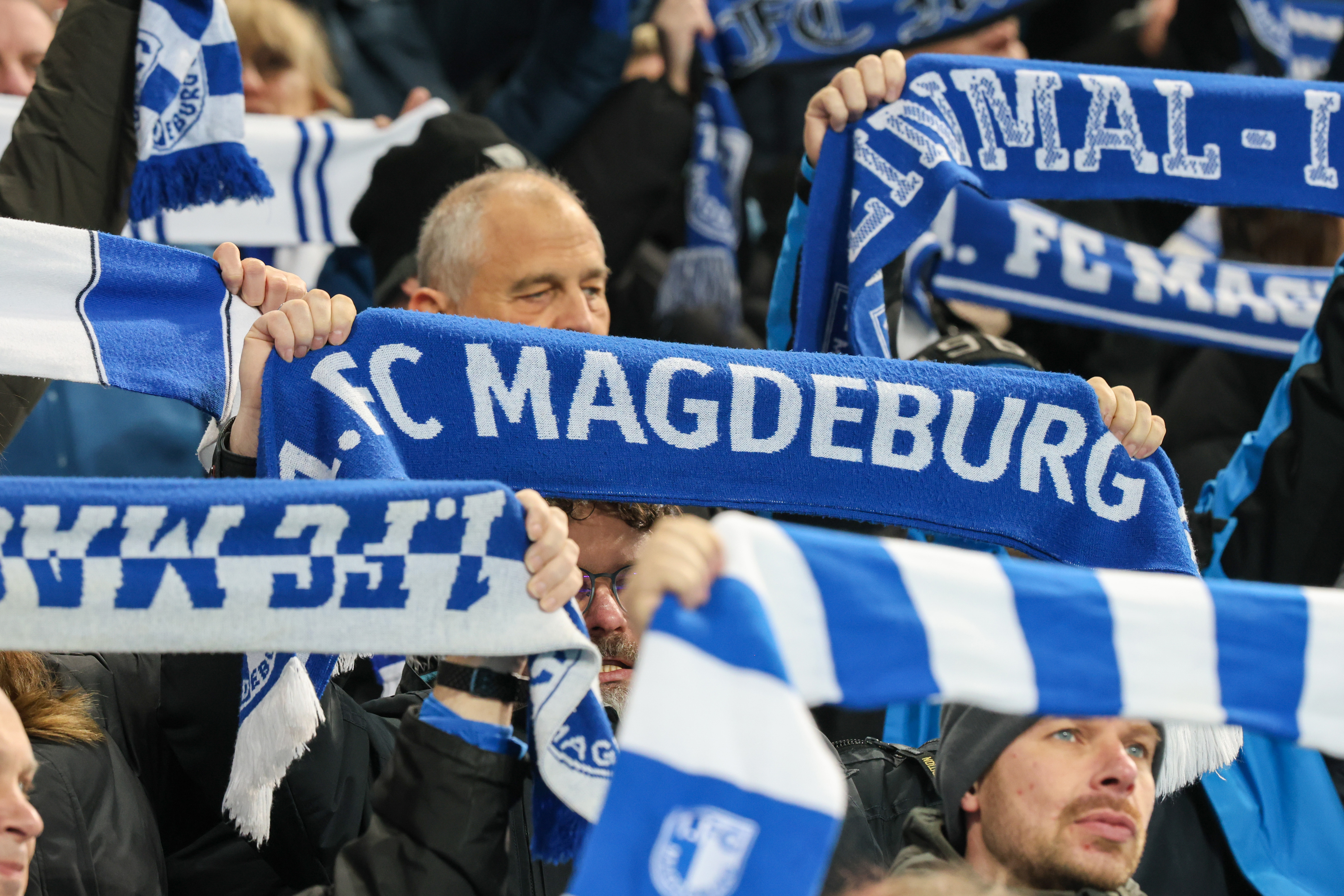 Fans des FCM stehen mit erhobenen blau weißen Schals im Stadion.