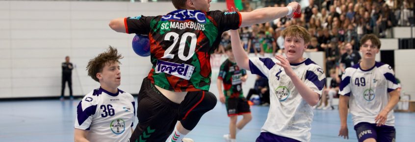 Ein Spieler der B-Jugend des SC Magdeburg (Trikotnummer 20) springt mit ausgestrecktem Arm zum Torwurf in einem Handballspiel gegen den TSV Bayer Dormagen. Zwei Gegenspieler in weißen Trikots mit blauen Elementen (Nummer 4 und 36) versuchen, den Wurf zu blocken. Im Hintergrund sind weitere Spieler, Zuschauer*innen und Tribünen zu sehen. Die Szene spielt in einer Sporthalle mit vielen Fans.