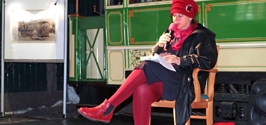 Anett Gröschner sitzt auf einem Stuhl vor einer historischen Straßenbahn und liest aus einem Buch vor. Sie ist rot und schwarz gekleidet.