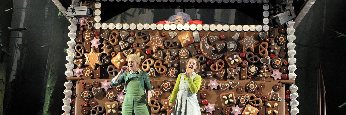 Zwei Darstellende auf heller Bühne, im Hintergrund ein buntes Lebkuchenhaus aus der Märchenoper Hänsel und Gretel.