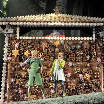 Zwei Darstellende auf heller Bühne, im Hintergrund ein buntes Lebkuchenhaus aus der Märchenoper Hänsel und Gretel.