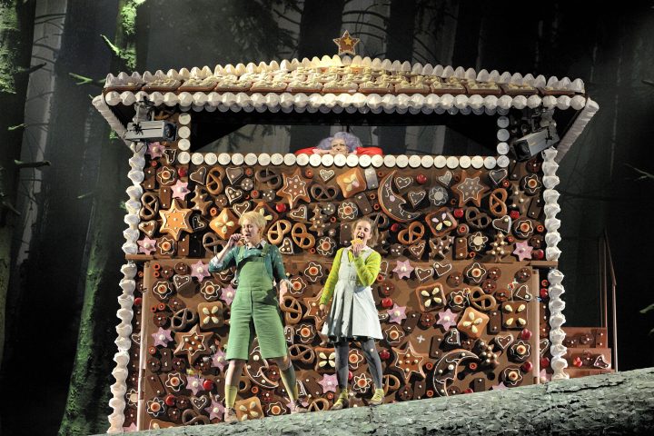 Zwei Darstellende auf heller Bühne, im Hintergrund ein buntes Lebkuchenhaus aus der Märchenoper Hänsel und Gretel.