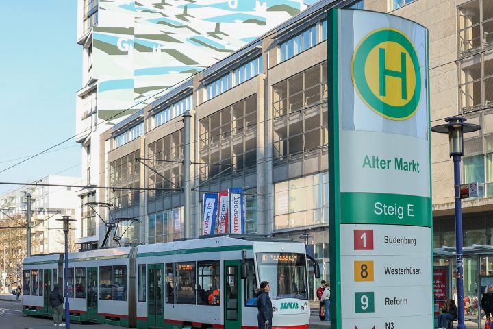 Im Vordergrund ist die Haltestellenstele der Haltestelle Alter Markt zu sehen und eine Straßenbahn steht an der Haltestelle.