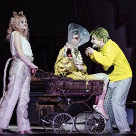 Bild aus der Oper Leben mit einem Idioten. Drei Personen sind auf der Bühne, eine sitzt in einem Kinderwagen. Sie sind grotesk gekleidet und geschminkt.