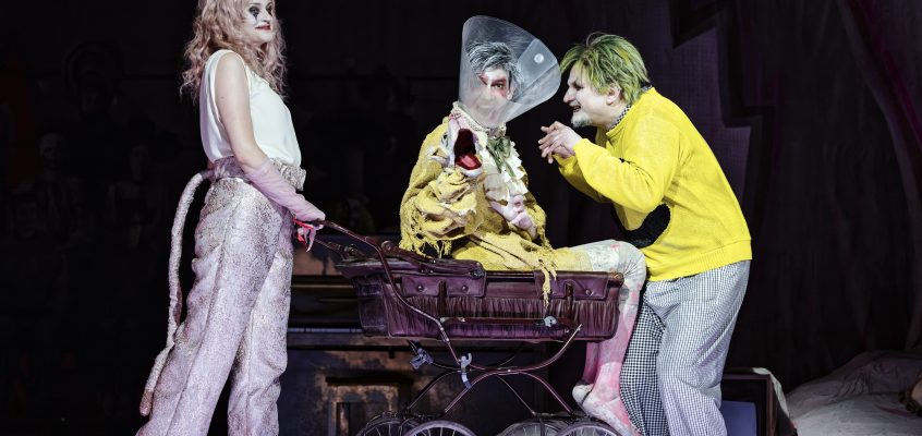 Bild aus der Oper Leben mit einem Idioten. Drei Personen sind auf der Bühne, eine sitzt in einem Kinderwagen. Sie sind grotesk gekleidet und geschminkt.