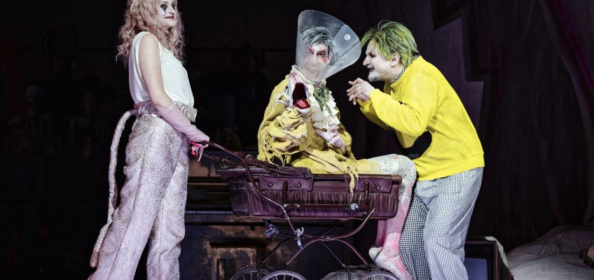 Bild aus der Oper Leben mit einem Idioten. Drei Personen sind auf der Bühne, eine sitzt in einem Kinderwagen. Sie sind grotesk gekleidet und geschminkt.