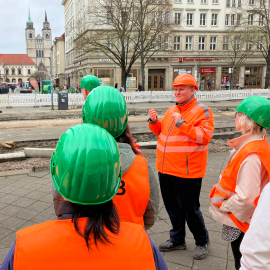 Es stehen mehrere Personen mit Schutzhelmen und Warnwesten bei einem geführten Baustellenspaziergang. Ein Mitarbeiter spricht zur Gruppe. Die Szene spielt auf der Baustelle am Breiten Weg in der Magdeburger Innenstadt.
