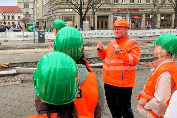 Es stehen mehrere Personen mit Schutzhelmen und Warnwesten bei einem geführten Baustellenspaziergang. Ein Mitarbeiter spricht zur Gruppe. Die Szene spielt auf der Baustelle am Breiten Weg in der Magdeburger Innenstadt.