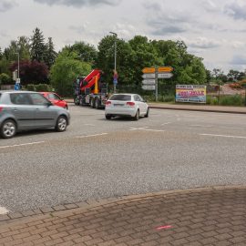 Die Aufnahme zeigt den Kreuzungsbereich an der Albert-Vater-Straße im Bereich der Abfahrt des Magdeburger Rings (MD-Ring). Im Vordergrund erstreckt sich eine mehrspurige Fahrbahn mit mehreren Fahrzeugen im fließenden Verkehr, darunter Pkw und ein Lkw mit Ladekran. Die Fahrbahn wirkt breit und stark frequentiert. Am rechten Fahrbahnrand befinden sich mehrere Wegweiser, die auf verschiedene Ziele im Stadtgebiet hinweisen. Dahinter ist eine eingezäunte Fläche mit Baustellen- bzw. Veranstaltungswerbung zu erkennen. Im Hintergrund prägen dichte Baum- und Grünflächen das Straßenbild. Links im Bild sind weitere Fahrspuren sowie die Zufahrt beziehungsweise Abfahrt des MD-Rings sichtbar. Der Himmel ist überwiegend bewölkt mit aufgelockerten Wolkenfeldern, wodurch eine leicht wechselhafte Wetterstimmung entsteht. Insgesamt vermittelt das Bild den Eindruck eines stark befahrenen Verkehrsknotens im urbanen Raum von Magdeburg.