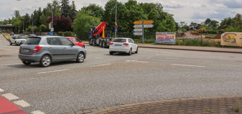 Die Aufnahme zeigt den Kreuzungsbereich an der Albert-Vater-Straße im Bereich der Abfahrt des Magdeburger Rings (MD-Ring). Im Vordergrund erstreckt sich eine mehrspurige Fahrbahn mit mehreren Fahrzeugen im fließenden Verkehr, darunter Pkw und ein Lkw mit Ladekran. Die Fahrbahn wirkt breit und stark frequentiert. Am rechten Fahrbahnrand befinden sich mehrere Wegweiser, die auf verschiedene Ziele im Stadtgebiet hinweisen. Dahinter ist eine eingezäunte Fläche mit Baustellen- bzw. Veranstaltungswerbung zu erkennen. Im Hintergrund prägen dichte Baum- und Grünflächen das Straßenbild. Links im Bild sind weitere Fahrspuren sowie die Zufahrt beziehungsweise Abfahrt des MD-Rings sichtbar. Der Himmel ist überwiegend bewölkt mit aufgelockerten Wolkenfeldern, wodurch eine leicht wechselhafte Wetterstimmung entsteht. Insgesamt vermittelt das Bild den Eindruck eines stark befahrenen Verkehrsknotens im urbanen Raum von Magdeburg.