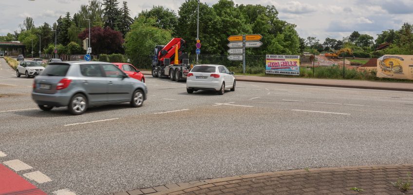 Die Aufnahme zeigt den Kreuzungsbereich an der Albert-Vater-Straße im Bereich der Abfahrt des Magdeburger Rings (MD-Ring). Im Vordergrund erstreckt sich eine mehrspurige Fahrbahn mit mehreren Fahrzeugen im fließenden Verkehr, darunter Pkw und ein Lkw mit Ladekran. Die Fahrbahn wirkt breit und stark frequentiert. Am rechten Fahrbahnrand befinden sich mehrere Wegweiser, die auf verschiedene Ziele im Stadtgebiet hinweisen. Dahinter ist eine eingezäunte Fläche mit Baustellen- bzw. Veranstaltungswerbung zu erkennen. Im Hintergrund prägen dichte Baum- und Grünflächen das Straßenbild. Links im Bild sind weitere Fahrspuren sowie die Zufahrt beziehungsweise Abfahrt des MD-Rings sichtbar. Der Himmel ist überwiegend bewölkt mit aufgelockerten Wolkenfeldern, wodurch eine leicht wechselhafte Wetterstimmung entsteht. Insgesamt vermittelt das Bild den Eindruck eines stark befahrenen Verkehrsknotens im urbanen Raum von Magdeburg.