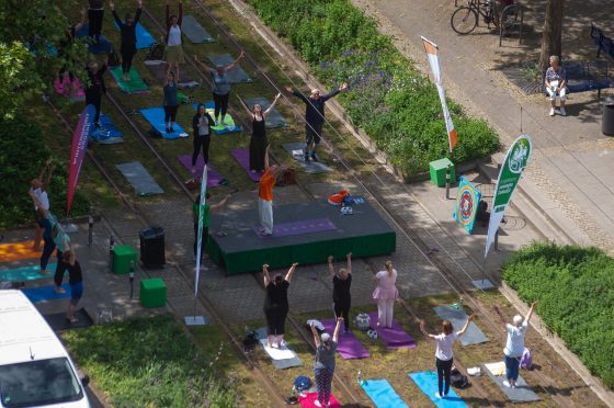 Luftaufnahme des Yoga Events auf dem Rasengleis des Breiten Wegs. An einem Fußgängerüberweg wurde eine Bühne aufgebaut, zu beiden Seiten der Bühne wurden Yogamatten auf dem Rasengleis ausgebreitet. Auf der Bühne steht eine Yogalehrerin, auf den Matten stehen die Kursmitglieder. Alle strecken ihre Arme nach oben.