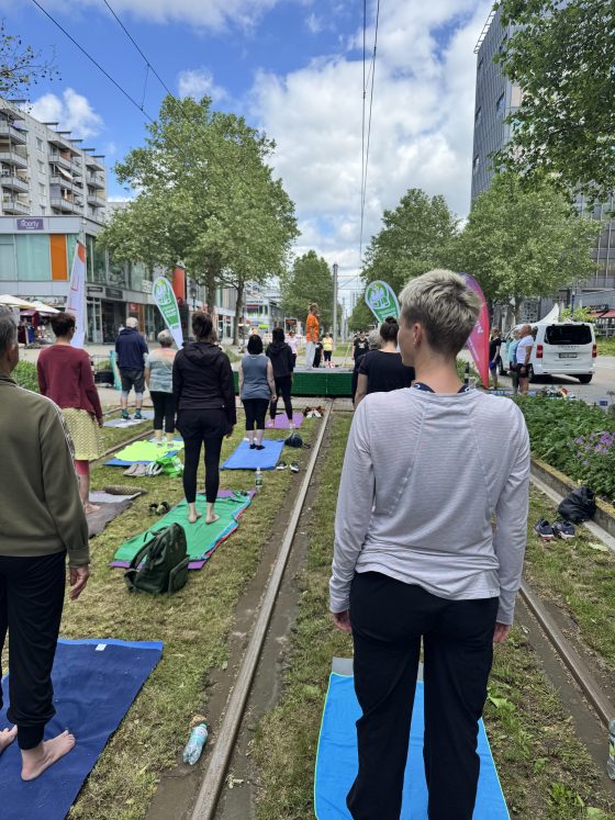 An einem Fußgängerüberweg wurde eine Bühne aufgebaut, zu beiden Seiten der Bühne wurden Yogamatten auf dem Rasengleis ausgebreitet. Auf der Bühne steht eine Yogalehrerin, auf den Matten stehen die Kursmitglieder.