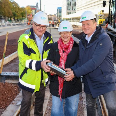 Andreas Fedorczuk (SWM), Birgit Münster-Rendel (MVB) und Jörg Rehbaum (Stadt Magdeburg) halten die Zeitkapsel auf der Baustelle Zukunftsgleise Innenstadt / Breiter Weg.